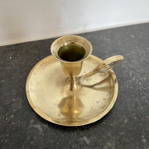 May include: A brass candlestick holder with a circular base and a small handle. The holder has a cup-shaped top to hold a candle. The brass has a warm, golden colour, and the design is simple and classic.