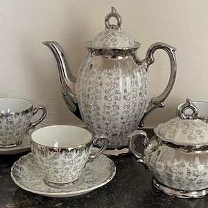 May include: A tea set featuring a teapot, a creamer, a sugar bowl with a lid, and multiple cups with matching saucers. Each piece is white porcelain with a metallic silver floral pattern and a reflective silver finish on the handles and rims.