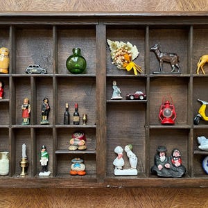 May include: A wooden printer's tray filled with various miniature figurines and objects. Includes small vases, bottles, figurines of people, animals, and vehicles, and other decorative items. The tray is brown and has many small compartments.