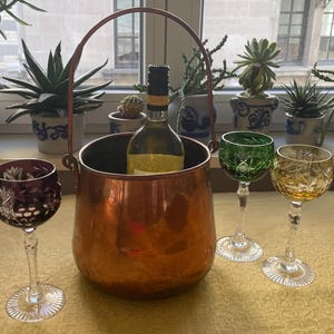 May include: A copper wine bucket with a bottle of wine inside, next to three crystal wine glasses in shades of purple, green, and yellow. The glasses have intricate cut patterns. The scene is set on a table near a window with potted plants.