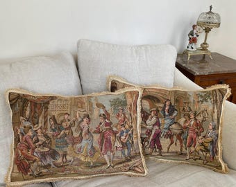Op de afbeelding: Twee decoratieve tapijtkussens met een beige franje. De kussens tonen een gedetailleerde scène van mensen die dansen en paardrijden in een Europese setting. Het kleurenpalet omvat rood, blauw en beige.
