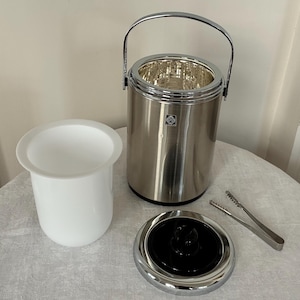 Può includere: Un secchiello per il ghiaccio cilindrico in acciaio inossidabile con manico basculante cromato, un contenitore interno rimovibile in plastica bianca, un coperchio cromato abbinato con pomello centrale nero e una pinza in metallo. Logo visibile.