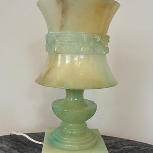 May include: A pale green and cream-colored stone table lamp with a flared, vase-shaped shade and a square base. The lamp features carved floral details around the center of the shade and the pedestal, with a white electrical cord extending from the side.