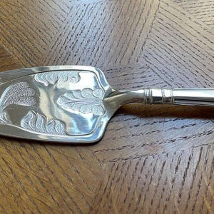 Op de afbeelding: Zilveren taartschep met een bloemmotief op het lemmet en de handgreep. De schep heeft een driehoekige lemmetvorm en een gedetailleerde, sierlijke handgreep. De schep is gemaakt van glanzend, reflecterend metaal.