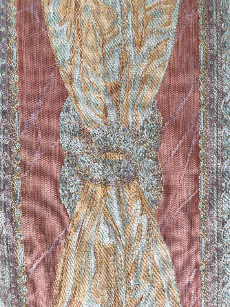 May include: Close-up of a textured fabric with a repeating pattern. The design features a central motif of intertwined shapes in shades of gold, green, and white, set against a reddish-brown background. The edges have a decorative border.