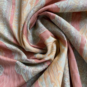 May include: Close-up of a draped fabric with a paisley pattern. The fabric features vertical stripes in shades of peach, pale orange, and cream. The fabric is soft and flowing, with intricate details in the design.