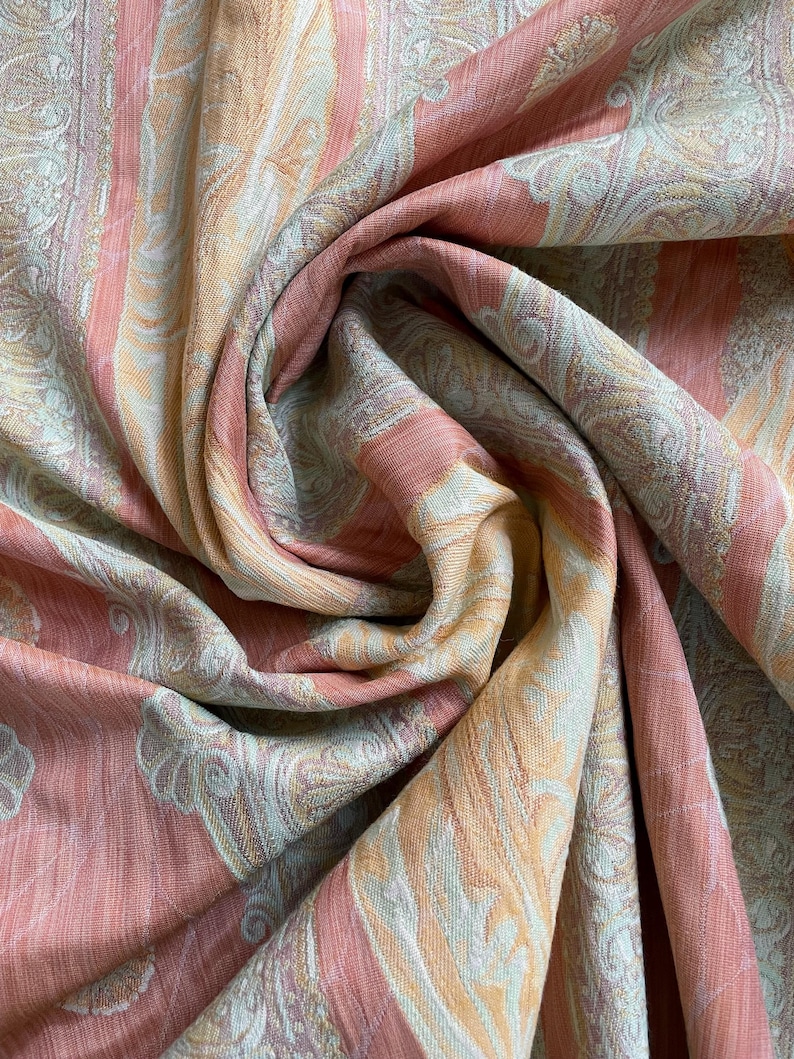 May include: Close-up of a draped fabric with a paisley pattern. The fabric features vertical stripes in shades of peach, pale orange, and cream. The fabric is soft and flowing, with intricate details in the design.
