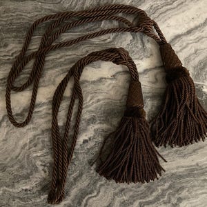 May include: Two brown decorative curtain tiebacks with tassels. The tiebacks are made of twisted rope and feature large, fluffy tassels at the ends. The tassels and rope are a rich, dark brown color.