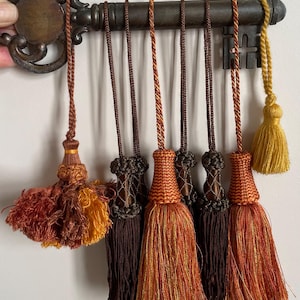 Pode incluir: Uma coleção de oito borlas decorativas em tons de castanho, laranja e amarelo, suspensas por uma grande chave de metal de estilo antigo. As borlas apresentam texturas variadas, incluindo cordões trançados, topos com nós e franjas soltas.