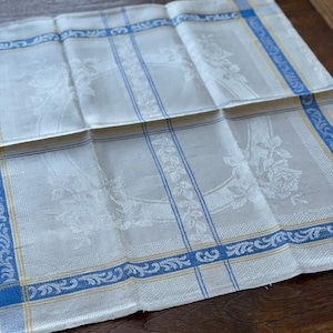 Pode incluir: Uma toalha de cozinha de linho branco com uma borda azul e amarela. A toalha tem um padrão floral e está dobrada ao meio.