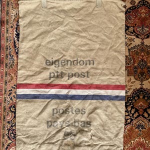 Puede incluir: Un saco de correo de lona vintage con rayas rojas, blancas y azules. El saco tiene el texto "eigendom pit post" y "postes pays-bas" impreso en él.