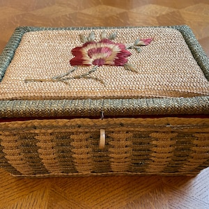 Op de afbeelding: Een rechthoekige geweven mand met een scharnierend deksel. Het deksel is versierd met een geborduurde bloem in rood- en roze tinten. De mand heeft een geweven ontwerp in beige en groen, met een groene rand. Ongeveer 25 cm lang.