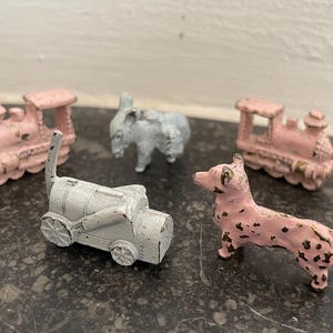 Może przedstawiać: Kolekcja małych, zabytkowych metalowych zabawek. Zabawki obejmują dwa różowe pociągi, szary wagon kolejowy, szarego słonia oraz różowego i czarnego psa w kropki. Zabawki są ułożone na ciemnej, nakrapianej powierzchni.
