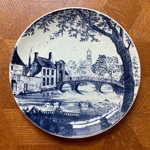 May include: A round, white and blue decorative plate with a landscape scene. The design depicts a bridge, buildings, trees, and swans on water. The plate has a dark blue border.