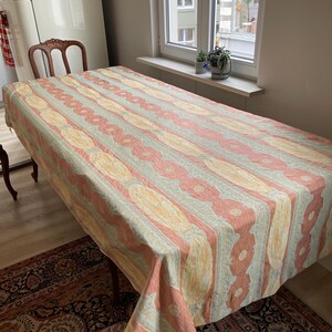 May include: A rectangular tablecloth with a repeating pattern of vertical stripes in peach, yellow, and gray. The tablecloth is draped over a wooden table, with a patterned rug visible beneath.
