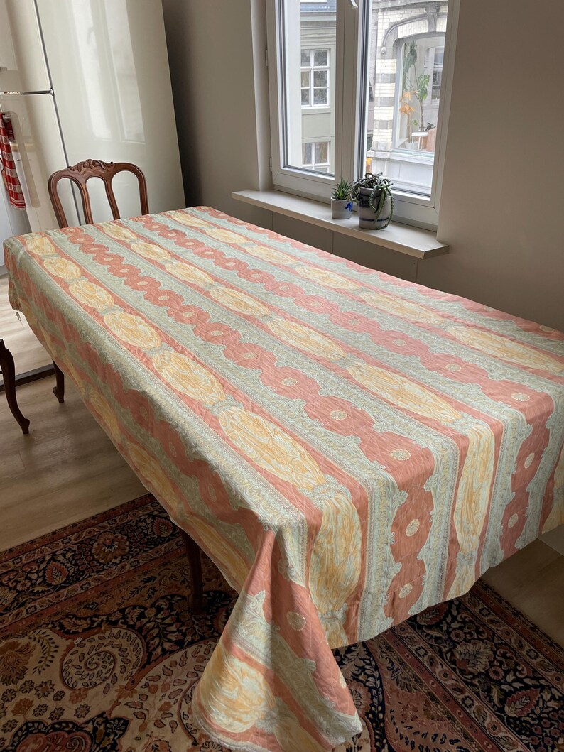 May include: A rectangular tablecloth with a repeating pattern of vertical stripes in peach, yellow, and gray. The tablecloth is draped over a wooden table, with a patterned rug visible beneath.