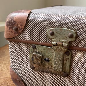 May include: A vintage suitcase featuring a textured beige and brown woven fabric exterior. The case includes weathered brown corner protectors and a tarnished metal latch mechanism with visible oxidation and a central keyhole.