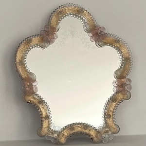 Puede incluir: Un espejo de pared con forma irregular y festoneada que presenta un borde texturizado en tono dorado. El marco está adornado con flores de vidrio transparente y rosa pálido en los bordes. La superficie central incluye un delicado diseño floral grabado.