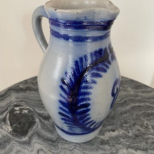 Puede incluir: Jarra de gres con cuerpo gris y asa azul. La jarra presenta bandas azules alrededor del cuello y un diseño azul en forma de helecho en la parte delantera. La jarra tiene un pico.