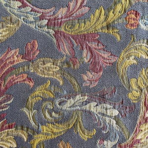 Könnte beinhalten: Nahaufnahme eines gewebten Stoffes mit detailliertem Blumenmuster. Das Design zeigt stilisierte Blätter und Schriftrollen in Gelb-, Rot- und Blautönen vor einem grauen Hintergrund. Der Stoff hat ein strukturiertes Aussehen, das auf einen Wandteppich oder Polsterstoff hindeutet.