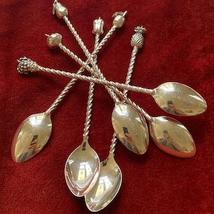 May include: A collection of silver spoons with twisted stems and fruit-shaped finials, including a pear, apple, and pineapple. The spoons are arranged on a red surface, showcasing their reflective surfaces and intricate designs. Each spoon has a small inscription.