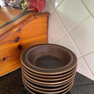 May include: A stack of ten brown ceramic bowls with a dark brown glaze. The bowls are stacked on a black surface.