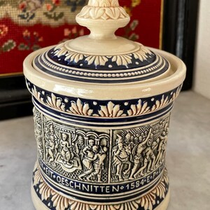 Könnte beinhalten: Eine Keramikdose mit Deckel in cremefarbener Ausführung, verziert mit dunkelblauen Details und Reliefkunst. Die Dose zeigt ein detailliertes Relief mit Figuren und dem Text "WEIT-OESCHNITTEN 1584-E-P". Der Deckel ist mit einer dekorativen Spitze versehen.