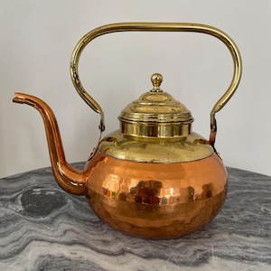 May include: A vintage copper and brass teapot with a hammered finish. The teapot has a rounded body, a curved spout, and a large brass handle. The lid is also brass and has a small knob on top. The teapot is sitting on a marble surface.