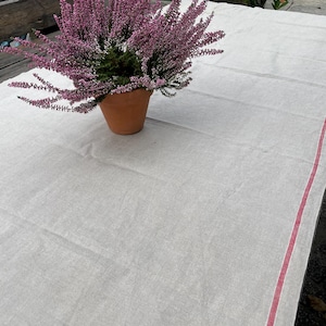 Pode incluir: Uma toalha de mesa de linho branco com uma faixa vermelha ao longo da borda. Uma planta em vaso com flores cor-de-rosa está centrada na toalha de mesa.