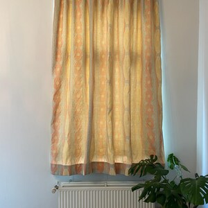 May include: A patterned curtain in shades of yellow, peach, and gray hangs in front of a window. The curtain features vertical stripes with a repeating floral design. The curtain is hanging from a white rod.
