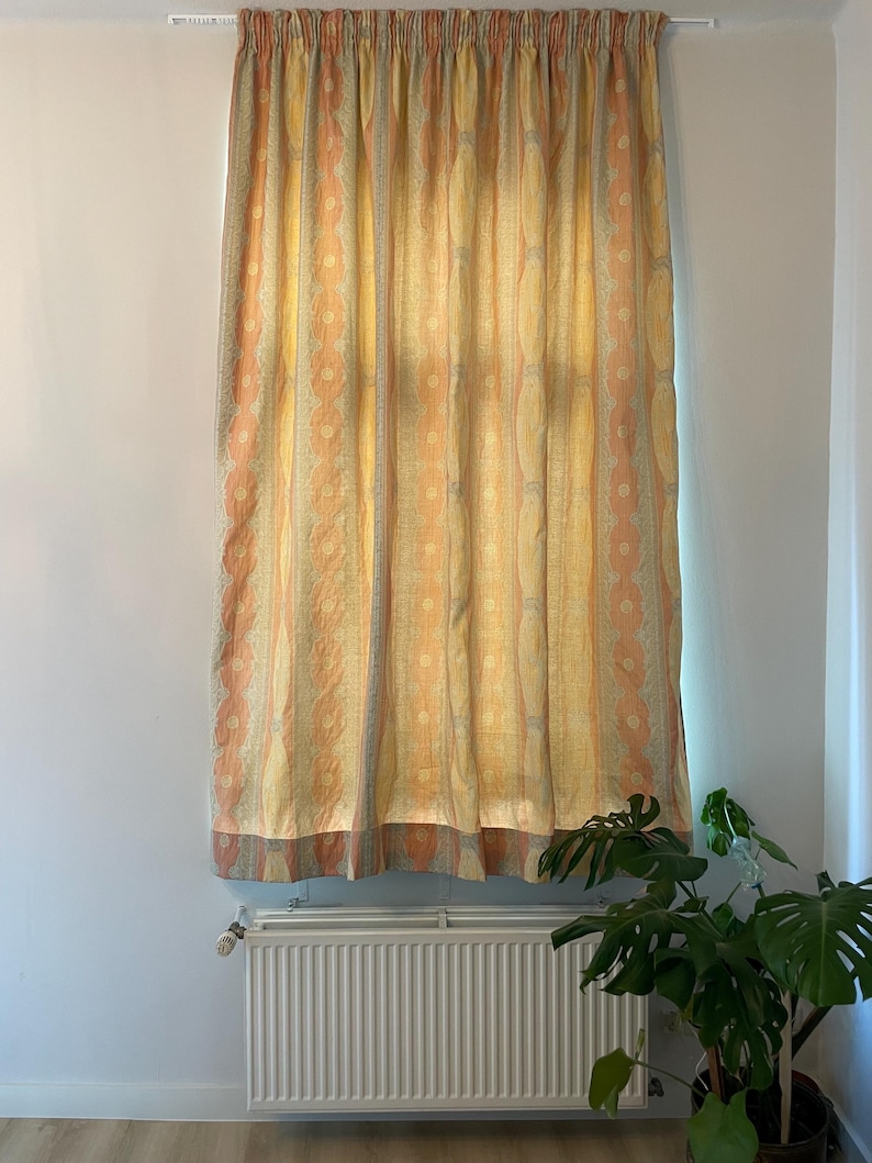 May include: A patterned curtain in shades of yellow, peach, and gray hangs in front of a window. The curtain features vertical stripes with a repeating floral design. The curtain is hanging from a white rod.