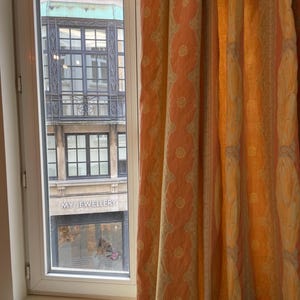May include: A window with a view of a building exterior and patterned curtains. The curtains are peach and yellow with a repeating circular design. The building has a green facade and the words "MY JEWELLERY" are visible.