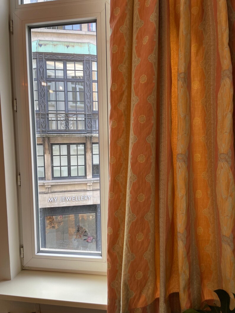 May include: A window with a view of a building exterior and patterned curtains. The curtains are peach and yellow with a repeating circular design. The building has a green facade and the words "MY JEWELLERY" are visible.