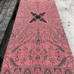 Può includere: Una grande coperta in lana con motivo paisley rosa con accenti neri e dorati. La coperta ha un bordo a frangia e un design centrale con una croce nera.