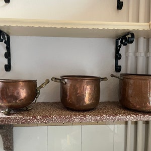 May include: Three copper pots with brass handles sit on a shelf. The pots are different sizes and have a hammered finish. The shelf is made of wood and has a white background.