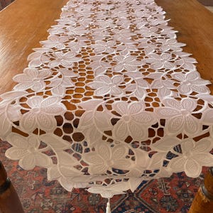 May include: White floral lace table runner. The runner is made of delicate, openwork lace with a repeating pattern of flowers and leaves. It is placed on a wooden table. A tassel hangs from the end of the runner.