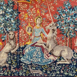 Può includere: Un arazzo tessuto raffigurante una figura centrale che regge uno specchio, affiancata da un leone e un unicorno su un'isola floreale blu scuro. Lo sfondo è un campo rosso vibrante pieno di piccoli fiori, alberi e animali, con frange dorate.