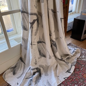 May include: Off-white linen curtain with a large-scale, abstract design in shades of gray. The pattern resembles brushstrokes or organic shapes. The curtain hangs in front of a window, with the bottom edge resting on a patterned rug.