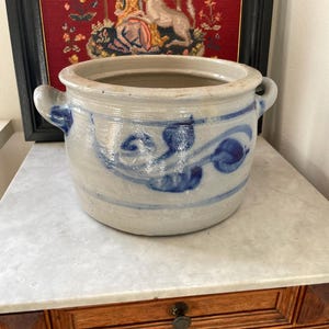 Puede incluir: Un cuenco de gres gris con diseños florales azules y dos asas. La olla está sobre una mesa de madera con tapa de mármol. Un tapiz enmarcado con un unicornio y figuras está en el fondo.