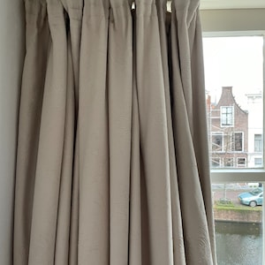 Op de afbeelding: Beige gordijnen met een textuurpatroon hangen aan een witte plafondrail. De gordijnen zijn dichtgetrokken en bedekken het zicht op een kanaal en gebouwen buiten het raam.