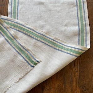 Pode incluir: Um corredor de mesa de linho branco com listras azuis, verdes e amarelas. O corredor está dobrado ao meio e tem uma borda desfiada.