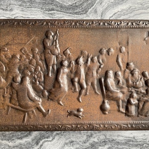 Op de afbeelding: Een vintage metalen plaquette met een reliëfdesign dat een groep mensen in een feestelijke scène voorstelt. De plaquette is bruin en heeft een decoratieve rand.