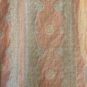 May include: Close-up of a peach-colored fabric with a quilted pattern. The design features vertical stripes and diamond shapes with floral and paisley-like motifs in cream and light gray. The fabric has a subtle sheen.