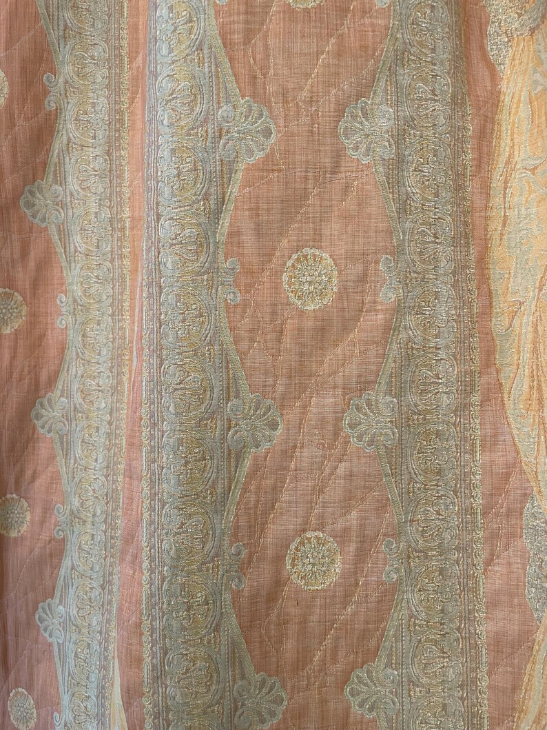 May include: Close-up of a peach-colored fabric with a quilted pattern. The design features vertical stripes and diamond shapes with floral and paisley-like motifs in cream and light gray. The fabric has a subtle sheen.