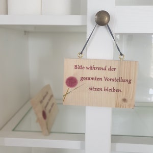 Könnte beinhalten: Ein Holzschild mit einer rosa Blumengrafik und dem Text "Bitte während der gesamten Vorstellung sitzen bleiben." hängt an einem weißen Regal.