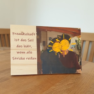 Könnte beinhalten: Ein Holzschild mit einem Zitat über Freundschaft: "Freundschaft ist das Seil, das hält, wenn alle Stränge reißen". Das Schild hat ein Foto von zwei Personen mit gelben sonnenförmigen Zeichnungen über ihren Gesichtern.