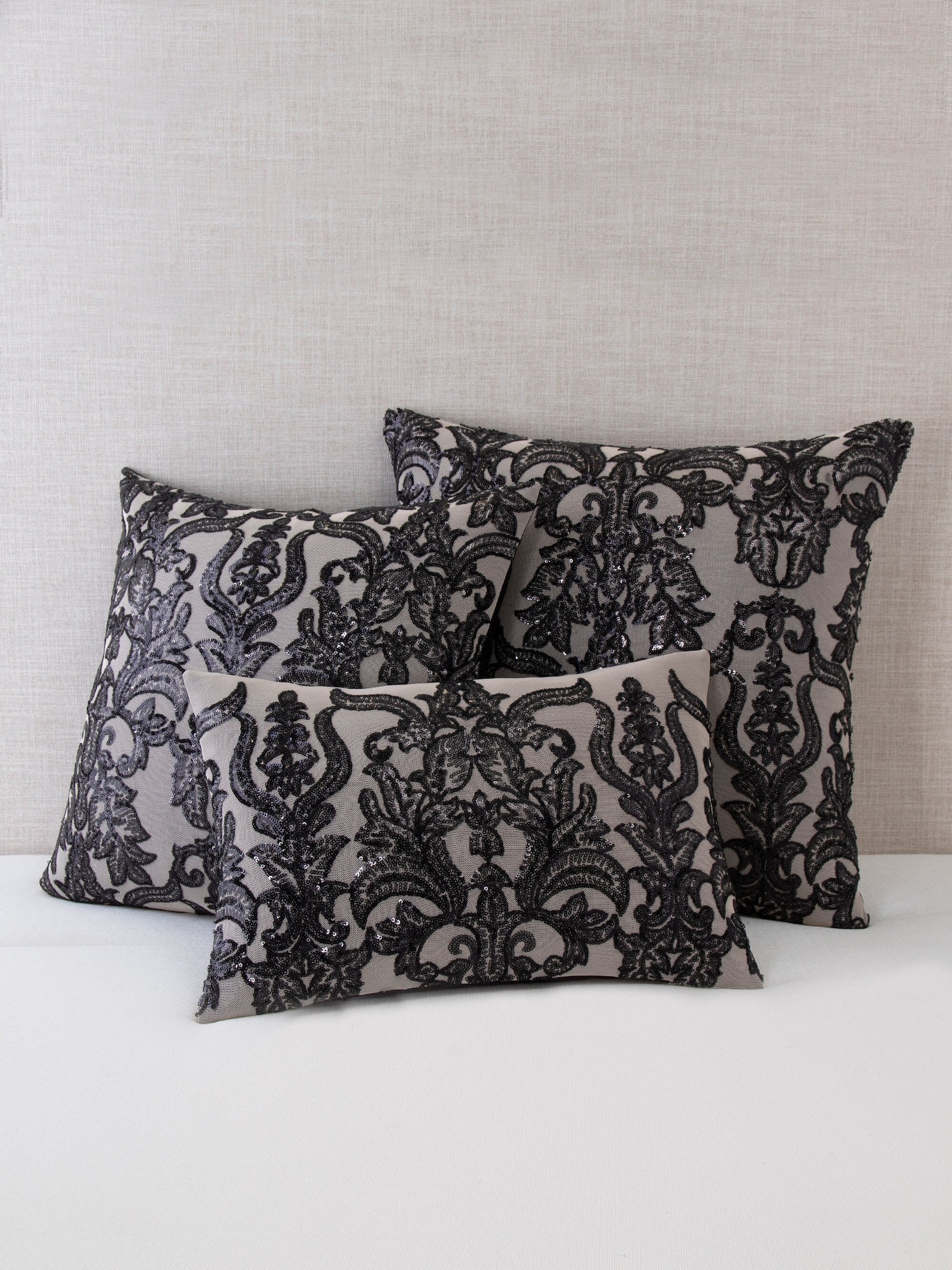 Set of three gray throw pillows with intricate black sequin damask embroidery, arranged on a white surface against a textured neutral backdrop