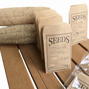 May include: A collection of seed packets and loofahs on a wooden surface. The seed packets are brown with the word "SEEDS" printed on them, along with handwritten details. The loofahs are beige and cylindrical.
