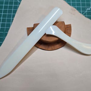 May include: Two white plastic tools for leather crafting. The larger tool is a long, flat blade with a rounded tip. The smaller tool is a curved blade with a rounded tip. Both tools are resting on a wooden base.