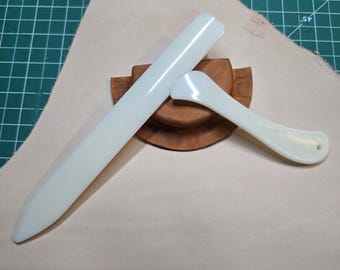 May include: Two white plastic tools for leather crafting. The larger tool is a long, flat blade with a rounded tip. The smaller tool is a curved blade with a rounded tip. Both tools are resting on a wooden base.
